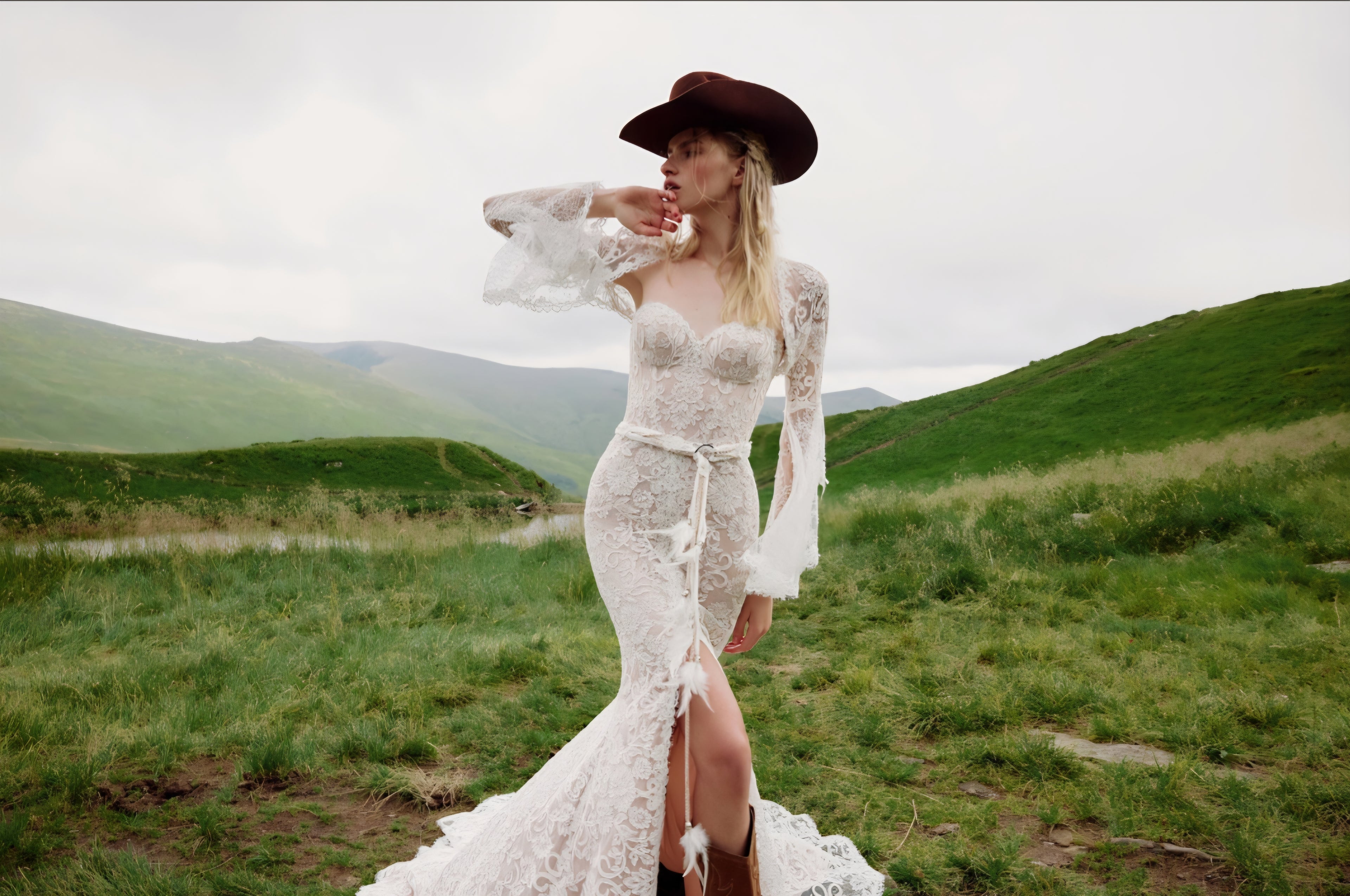 Woman in a white lace dress and brown hat standing in a grassy field with mountains in the background