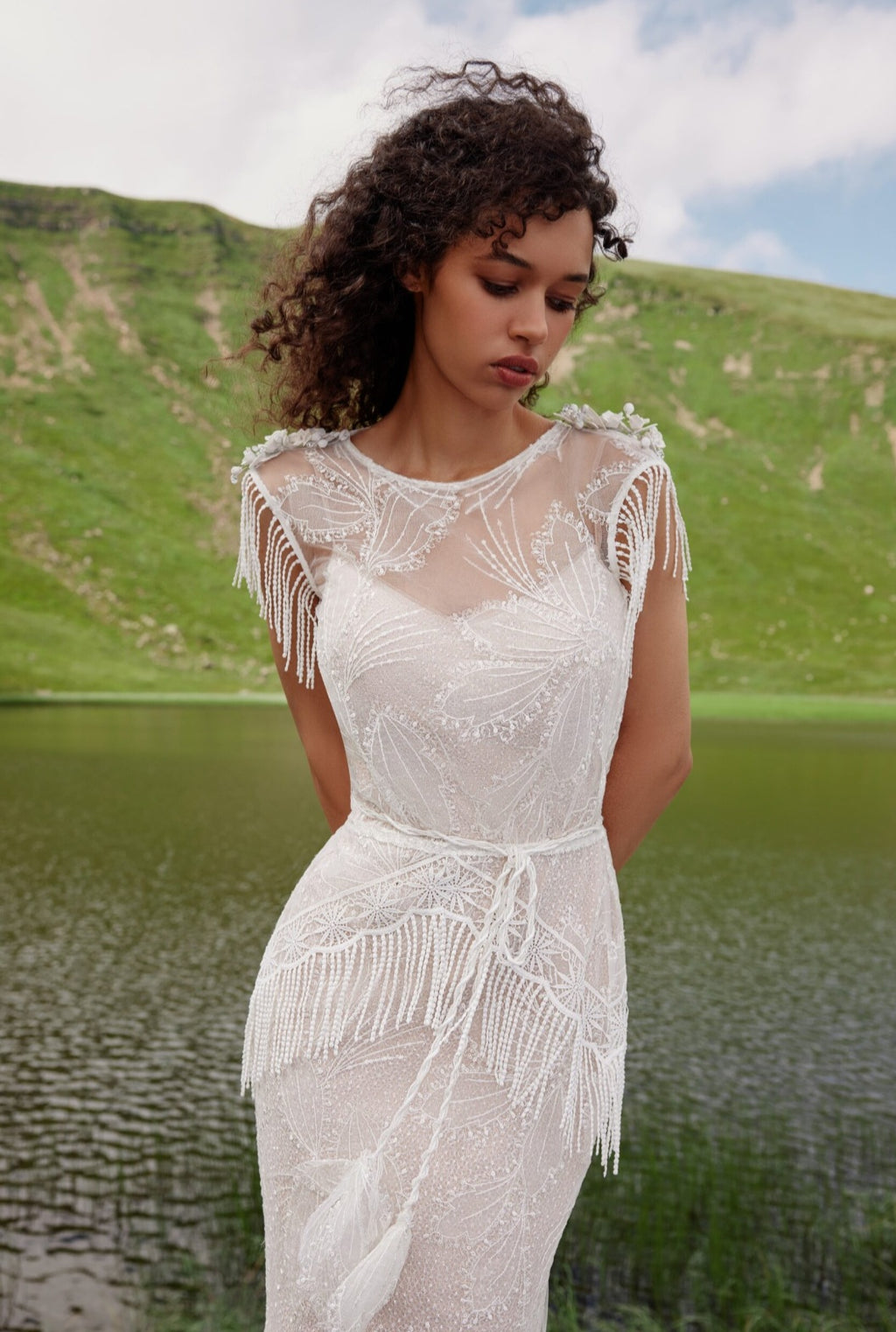 Woman in a white lace dress standing by a lake with green hills in the background