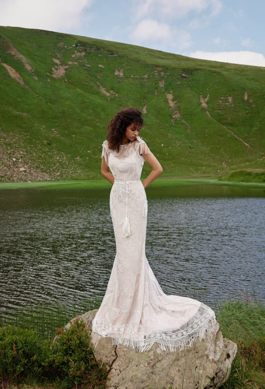 Woman in a white lace dress standing on a rock by a lake with green hills in the background