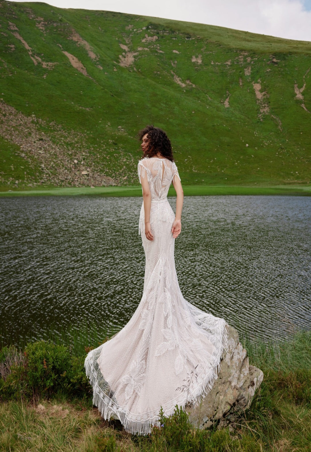 Woman in a white lace dress standing by a lake with green hills in the background