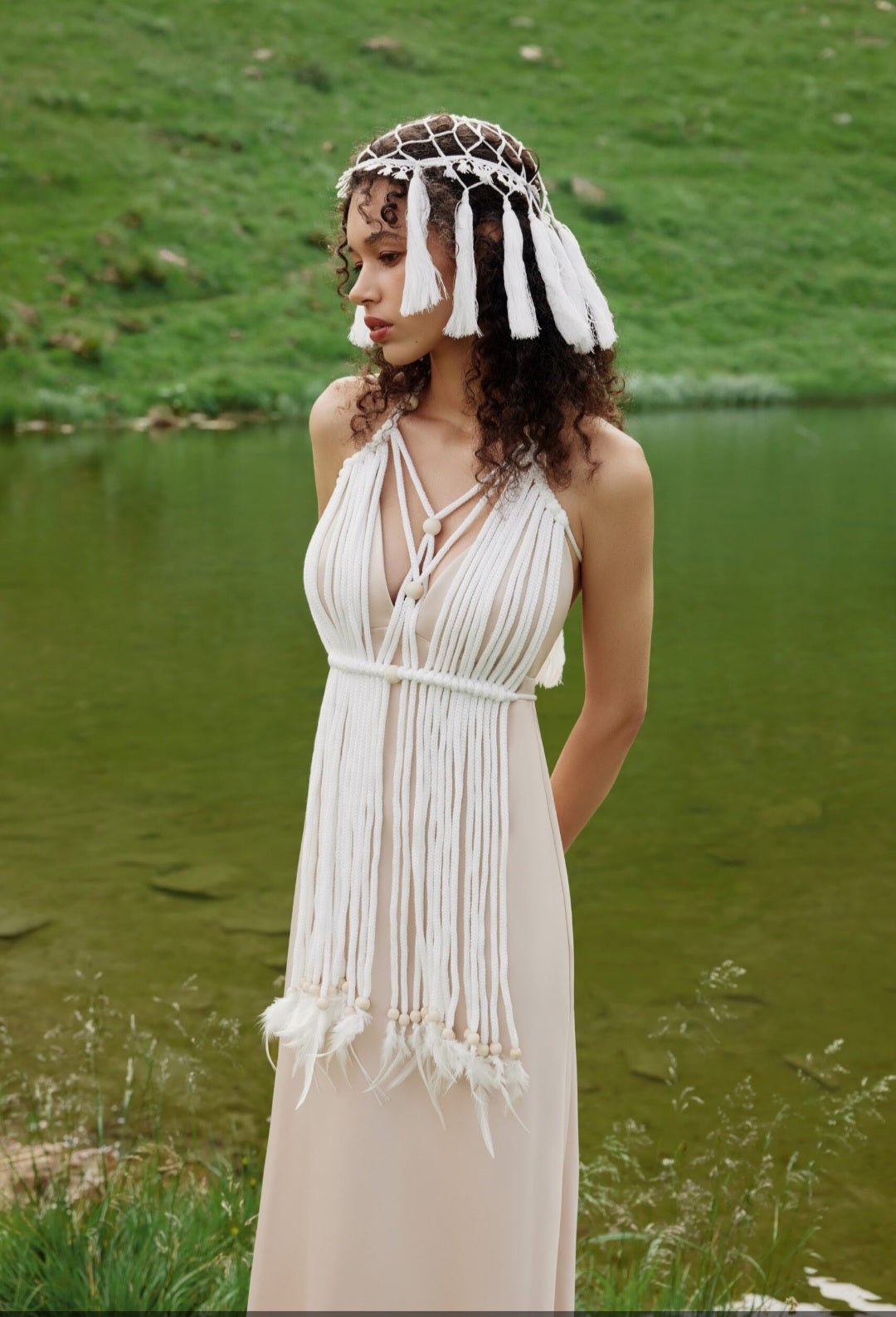 Woman in a flowing dress and headpiece standing by a body of water.