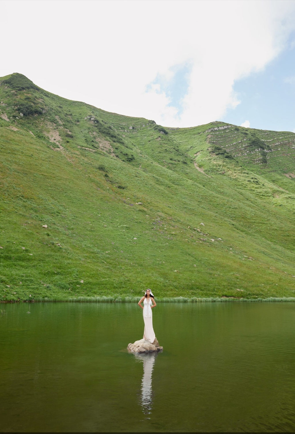 Person in a white dress standing on a rock in a lake with green mountains in the background