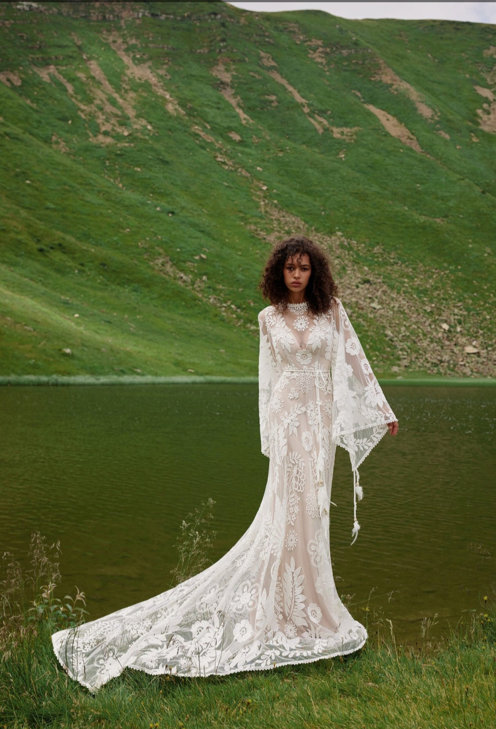 Woman in a white lace dress standing in a natural setting with green hills and water.