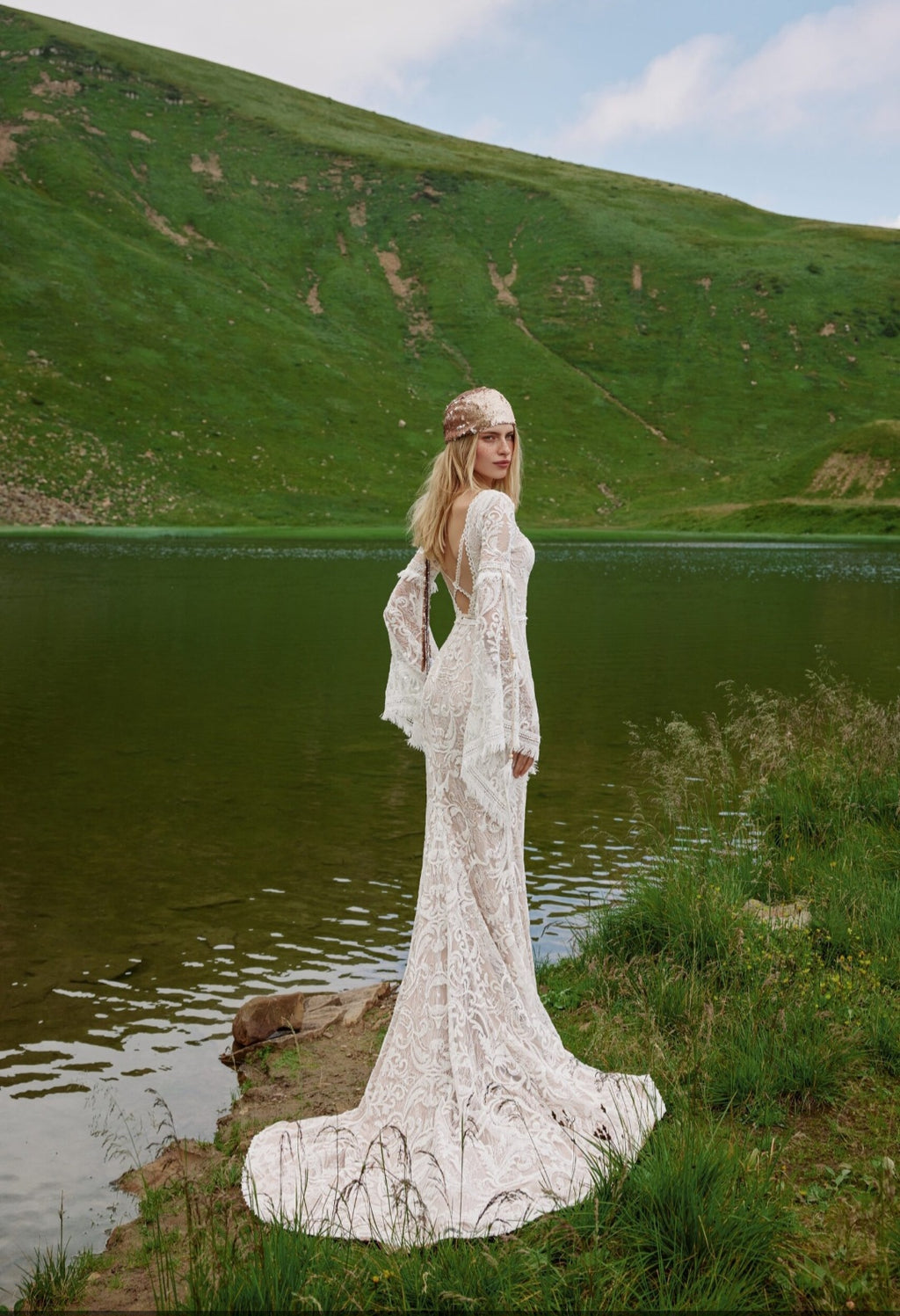 Woman in a white lace dress standing by a lake with green hills in the background