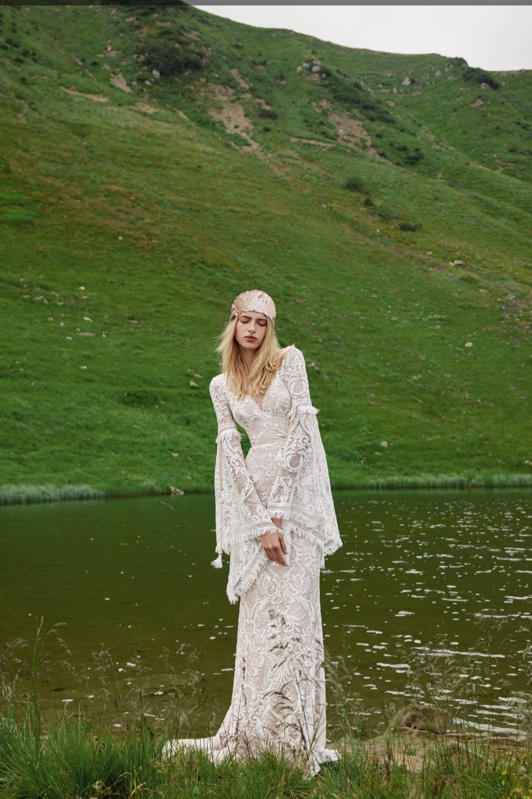 Woman in a white lace dress standing by a lake with green hills in the background