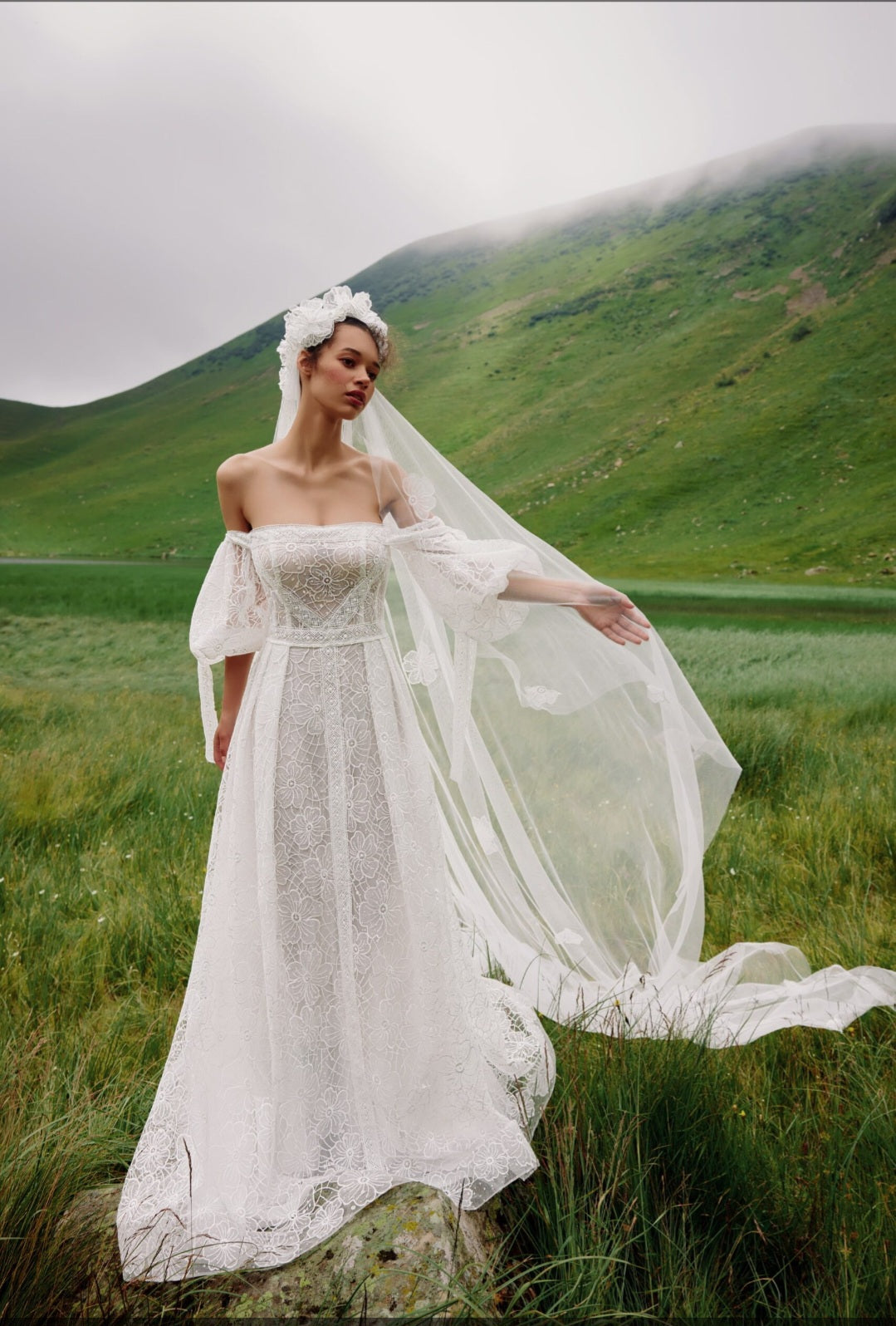 Woman in a white wedding dress standing in a grassy field with mountains in the background