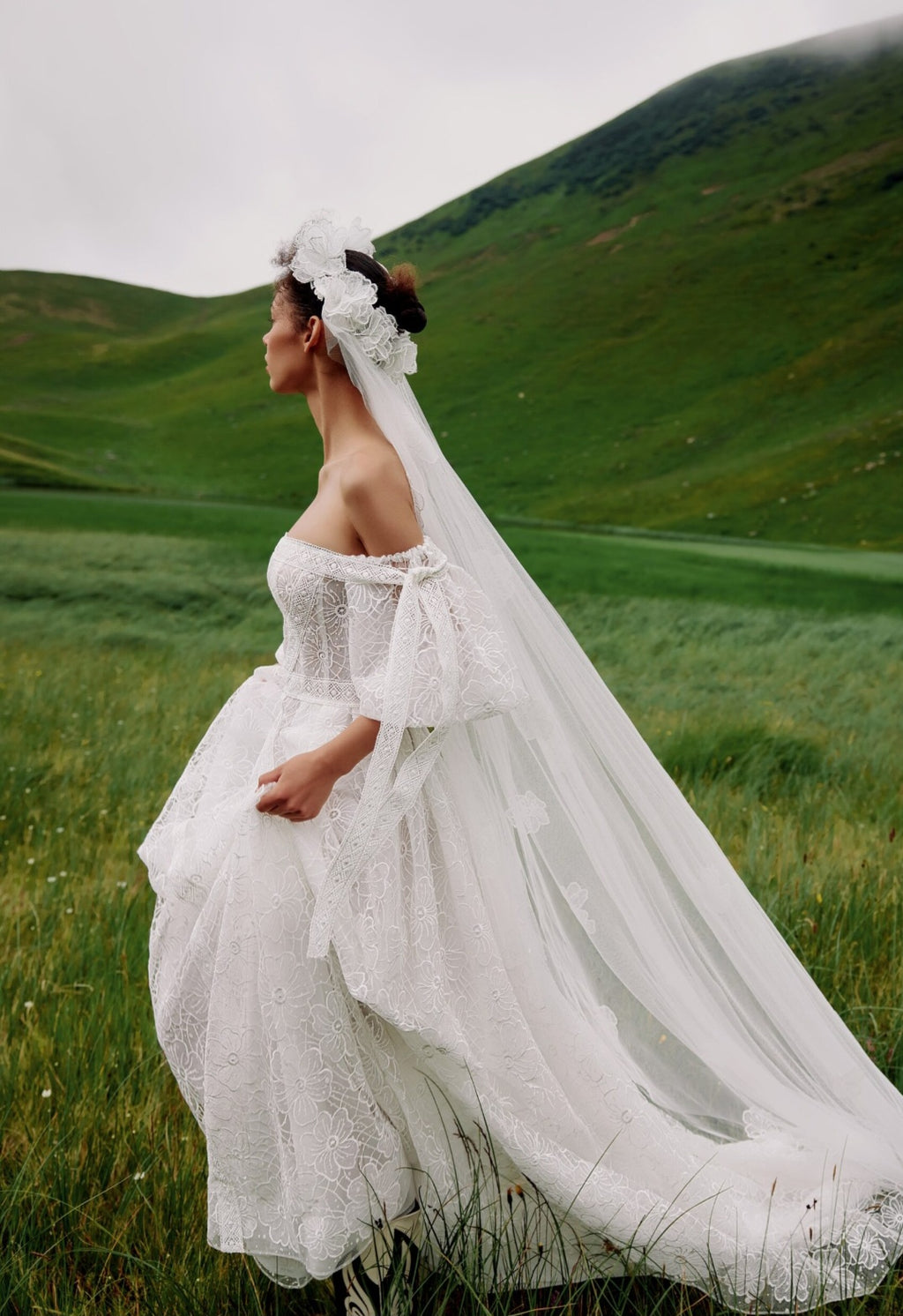 Woman in a white wedding dress with a long veil standing in a green field.