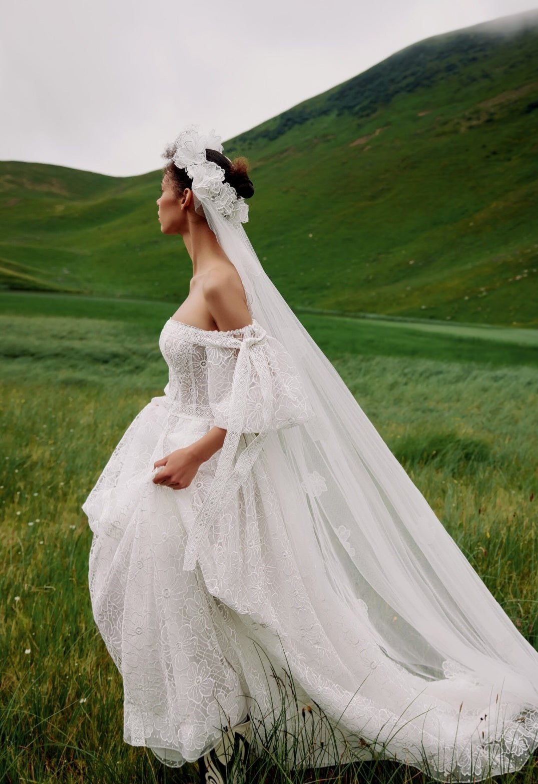 Woman in a white wedding dress with a long veil standing in a green field.