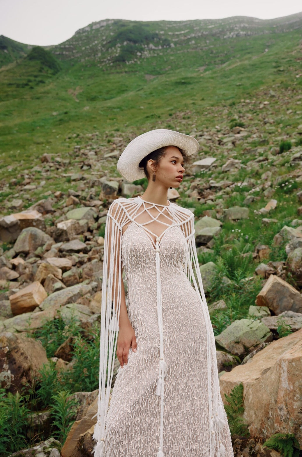 Woman in a white dress and hat standing in a rocky, mountainous landscape.