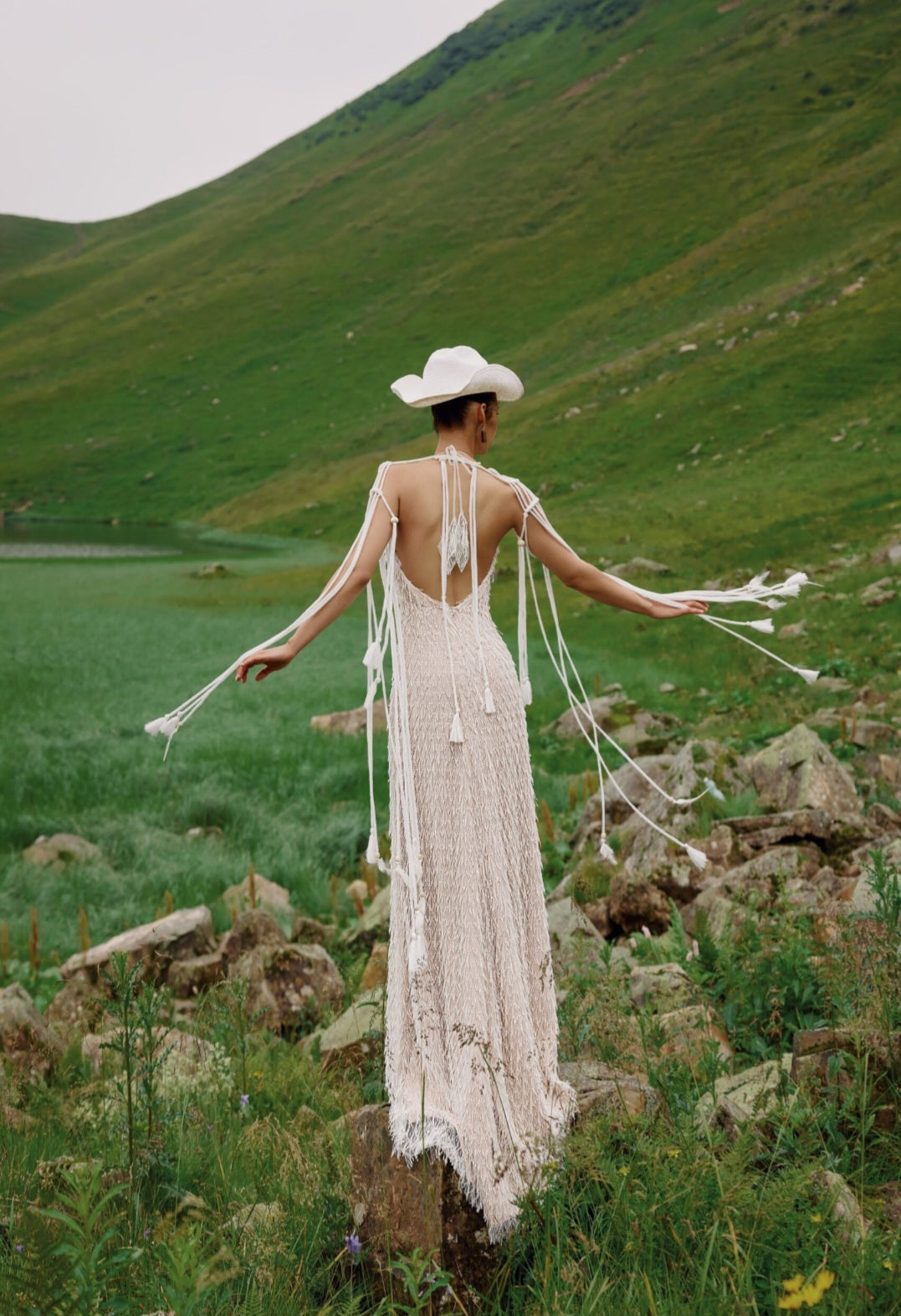 Person in a long, flowing dress with tassels standing in a mountainous landscape.