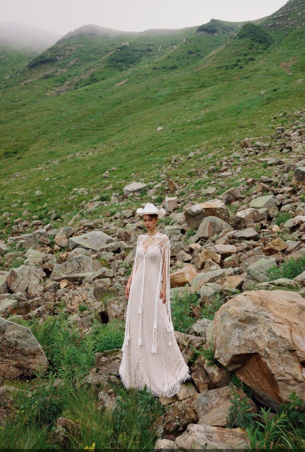 Woman in a white dress standing on rocky terrain with green mountains in the background
