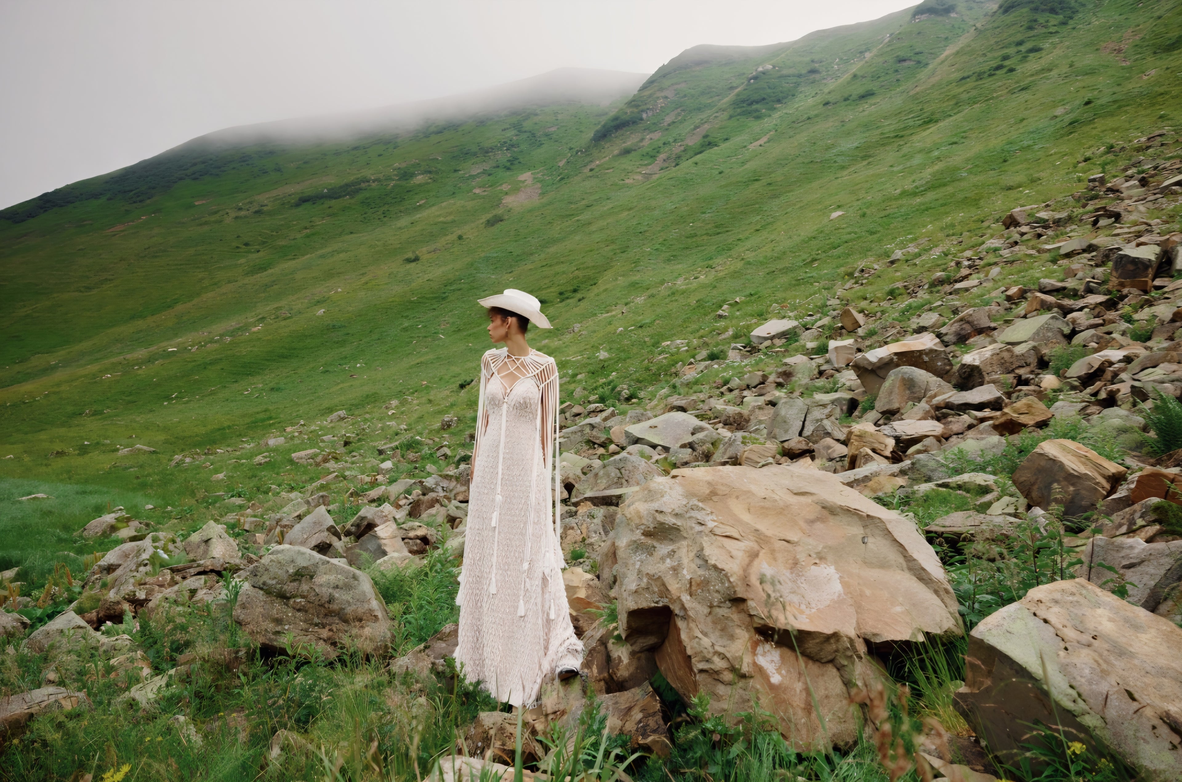 Person in a white dress standing on a rocky hillside with green valleys in the background