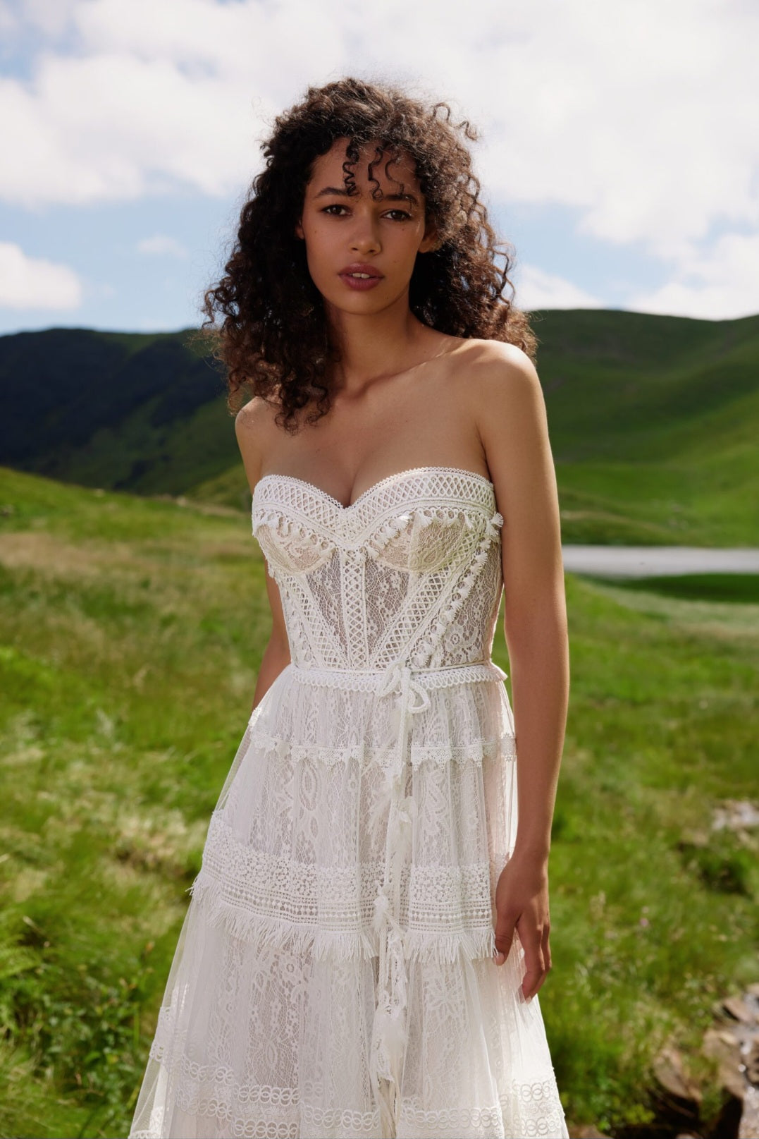 Woman in a white lace dress standing in a scenic outdoor setting with green hills and blue sky.