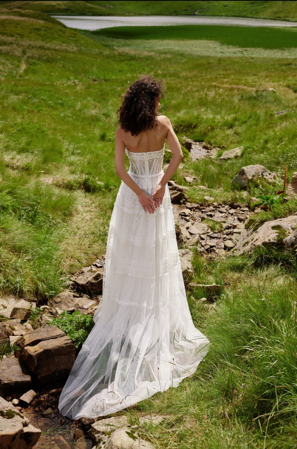 Woman in a white dress standing on a grassy hill with a pond in the background