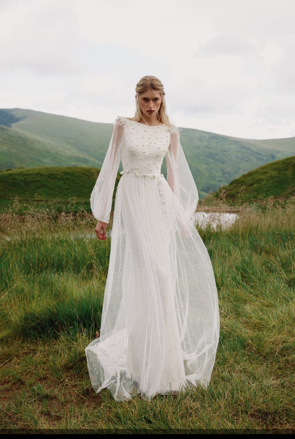 Woman in a white wedding dress standing in a grassy field with mountains in the background