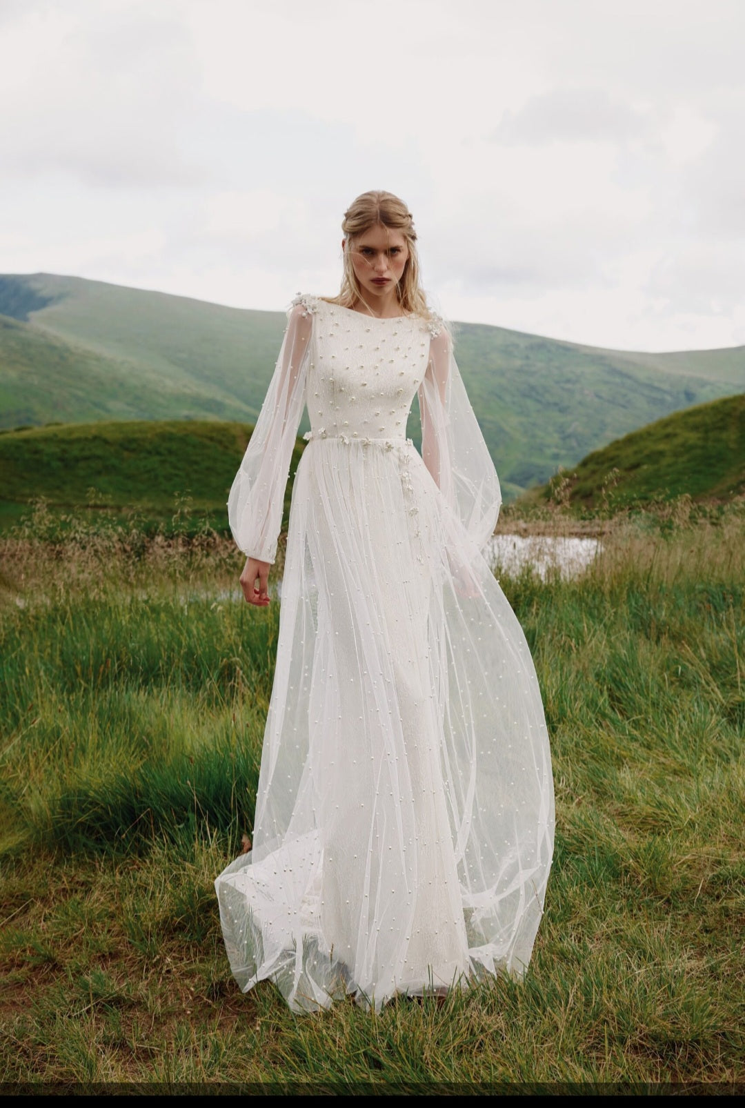 Woman in a white wedding dress standing in a grassy field with mountains in the background