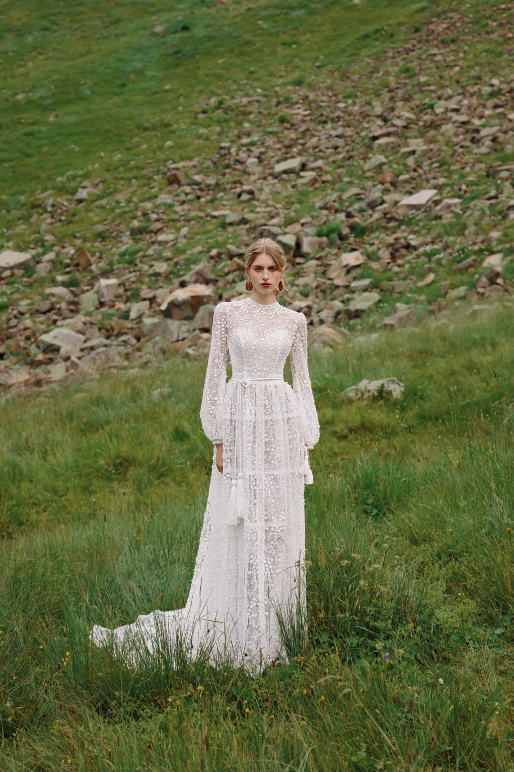 Woman in a white lace dress standing in a grassy field with a rocky hill in the background