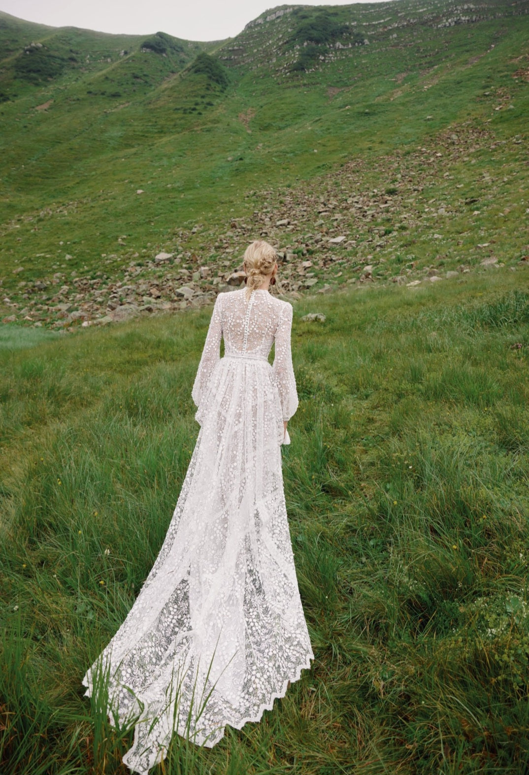 Person in a white lace dress standing in a grassy field with mountains in the background