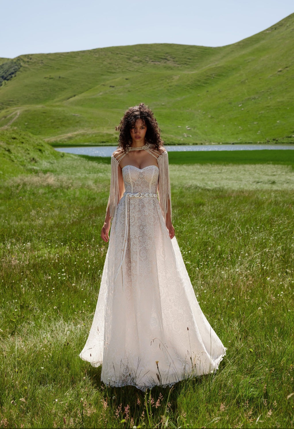 Woman in a white dress standing in a grassy field with a lake and hills in the background