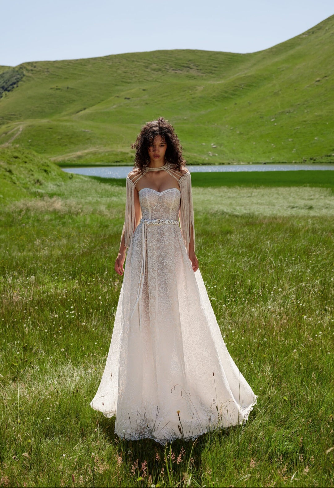 Woman in a white dress standing in a grassy field with a lake and hills in the background