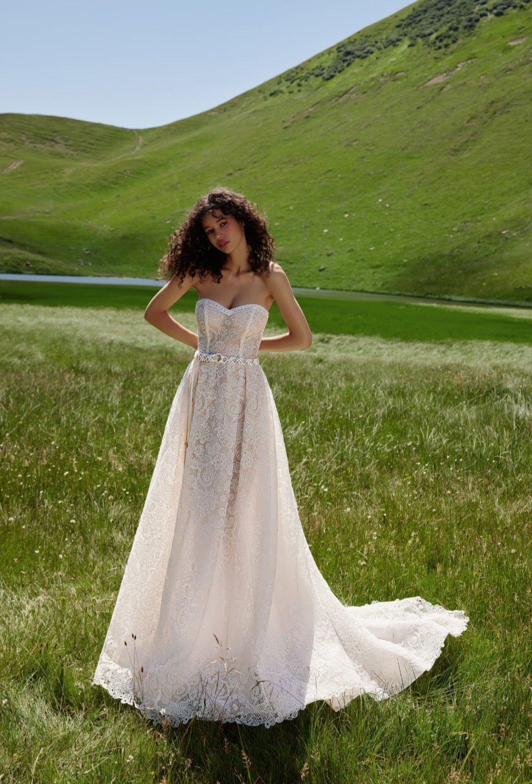 Woman in a white wedding dress standing in a grassy field with hills in the background