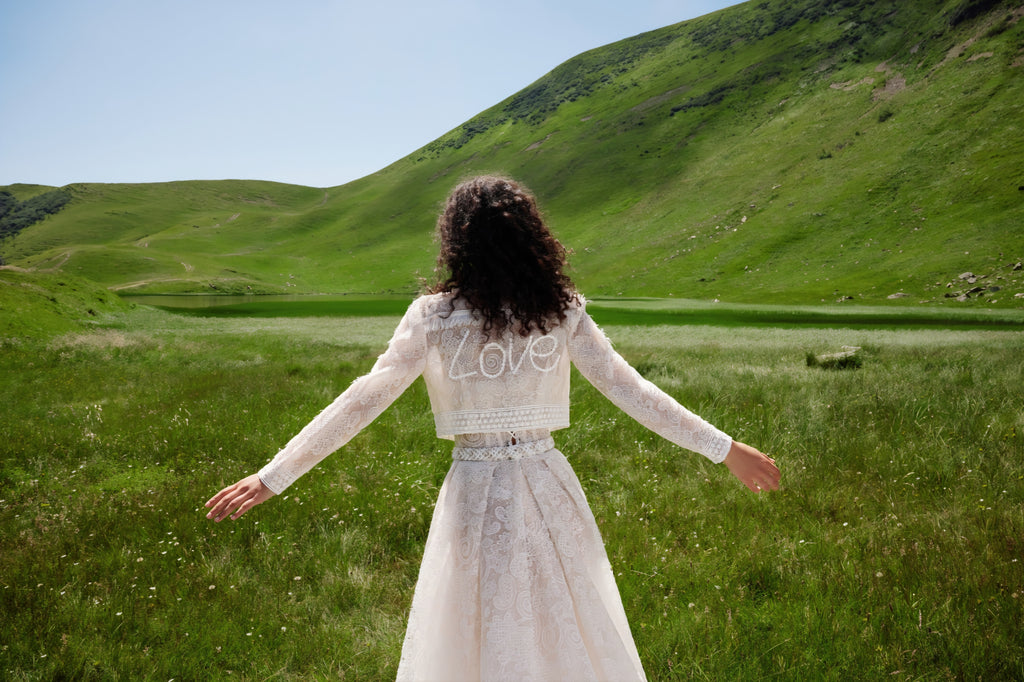 Woman in a white dress with 'LOVE' on her jacket standing in a green field with mountains in the background