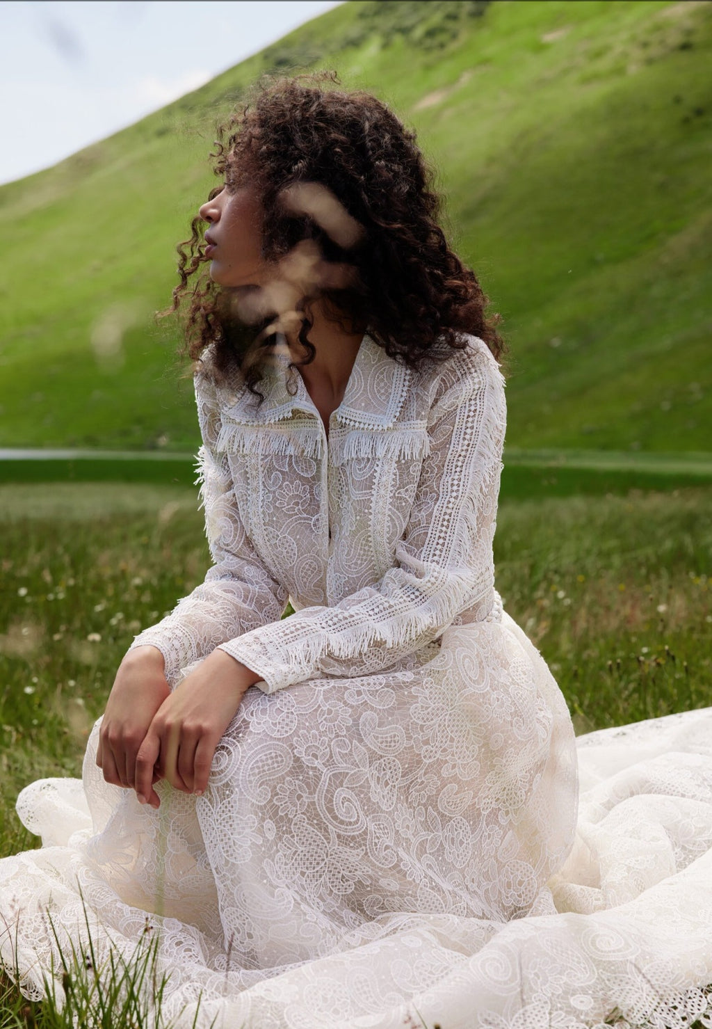 Woman in a white lace dress sitting in a grassy field with a dog on her lap.