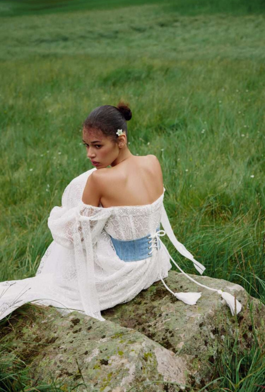 Woman in a white off-shoulder top and denim skirt sitting on a rock in a grassy field.