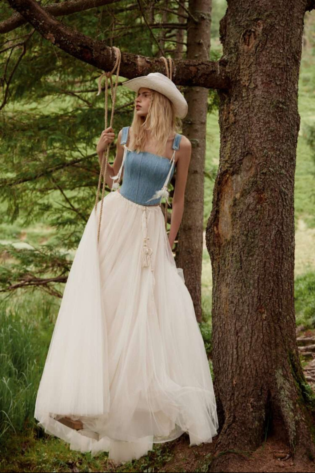 Woman in a blue top and white skirt standing next to a tree in a forest