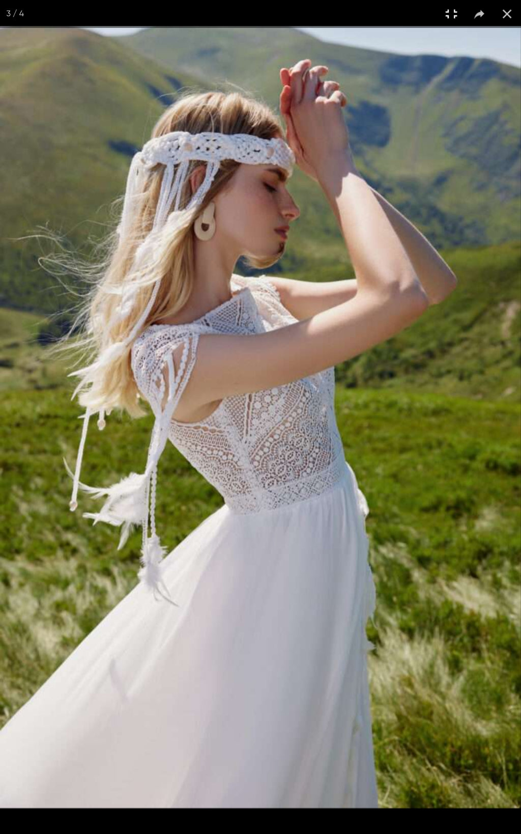 Woman in a white dress with lace details standing in a grassy field with mountains in the background