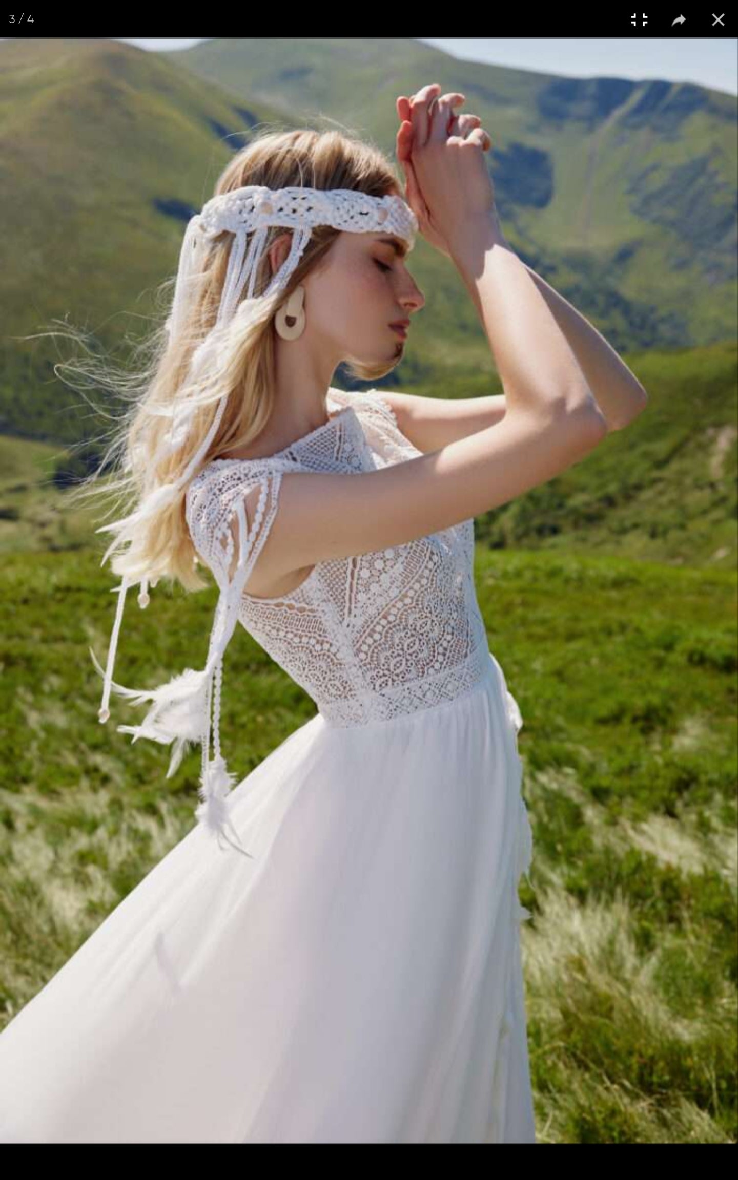 Woman in a white dress with lace details standing in a grassy field with mountains in the background