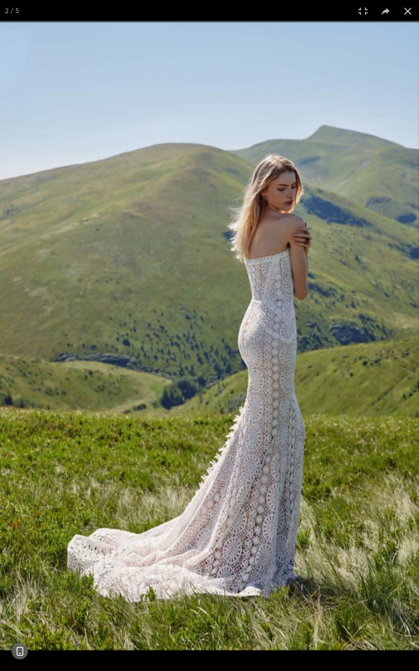 Woman in a white lace wedding dress standing in a grassy field with mountains in the background