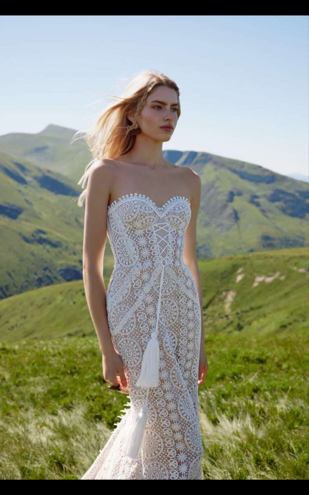 Woman in a white lace wedding dress standing in a grassy field with mountains in the background