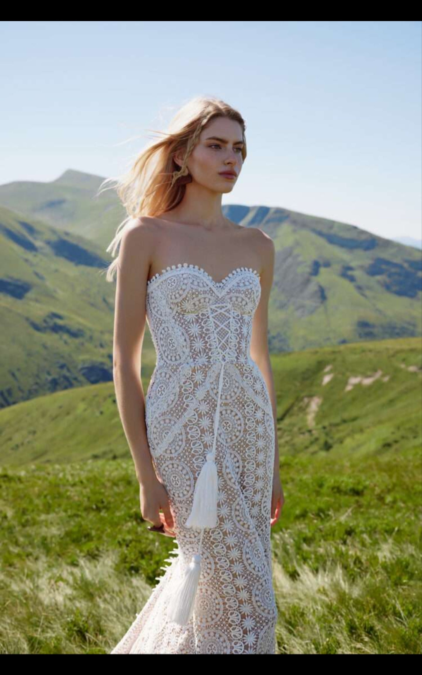 Woman in a white lace wedding dress standing in a grassy field with mountains in the background