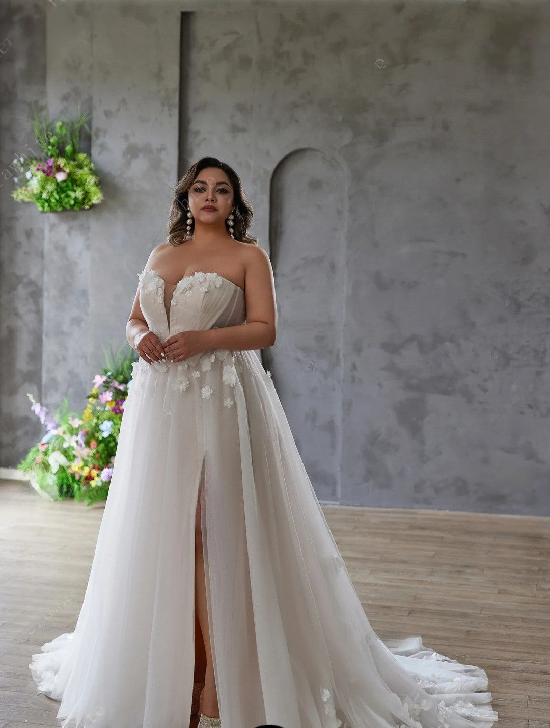Woman in a white strapless gown with floral details standing against a gray wall.