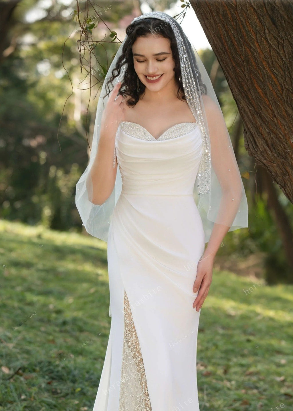 Woman in a white wedding dress with a veil standing outdoors near trees.