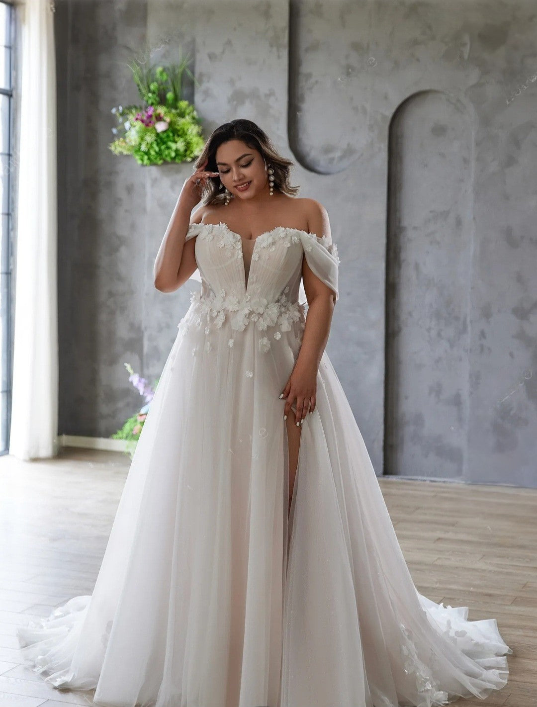 Woman in a white wedding dress with floral details standing in a room with a gray wall and plants.
