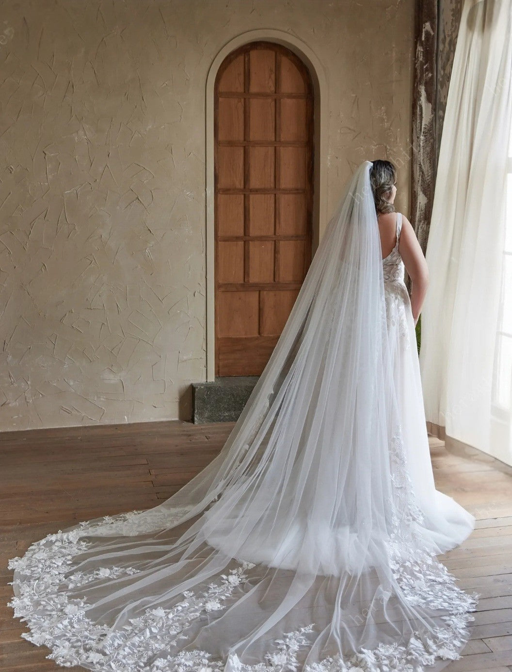 Bride in a wedding dress with a long veil standing in a room with wooden floor and door.