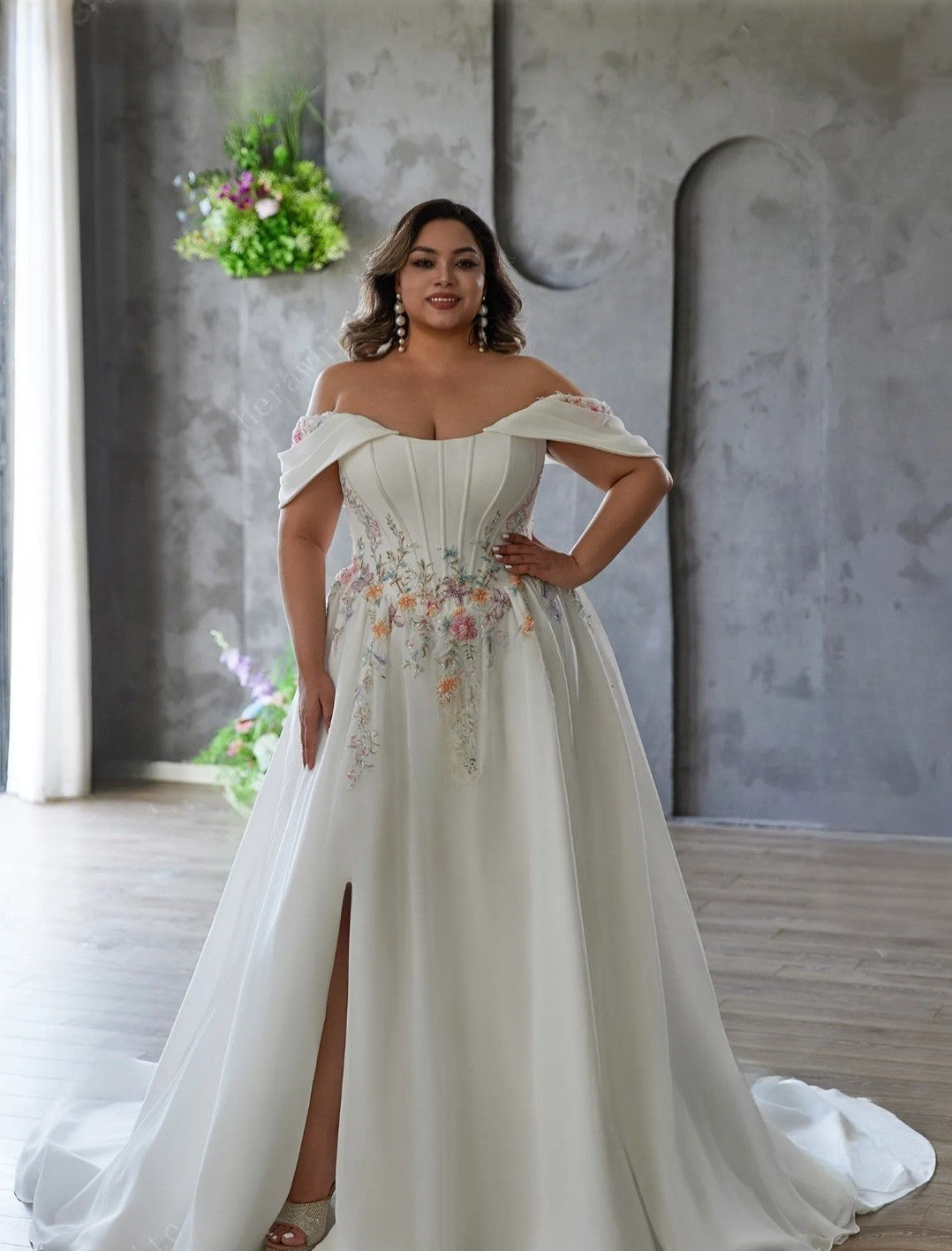 Woman in a white off-shoulder gown with floral embroidery standing in a modern interior setting.