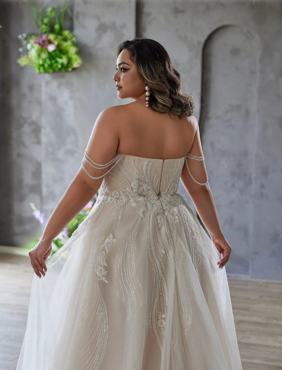 Woman in a white wedding dress with floral details standing in a softly blurred indoor setting.