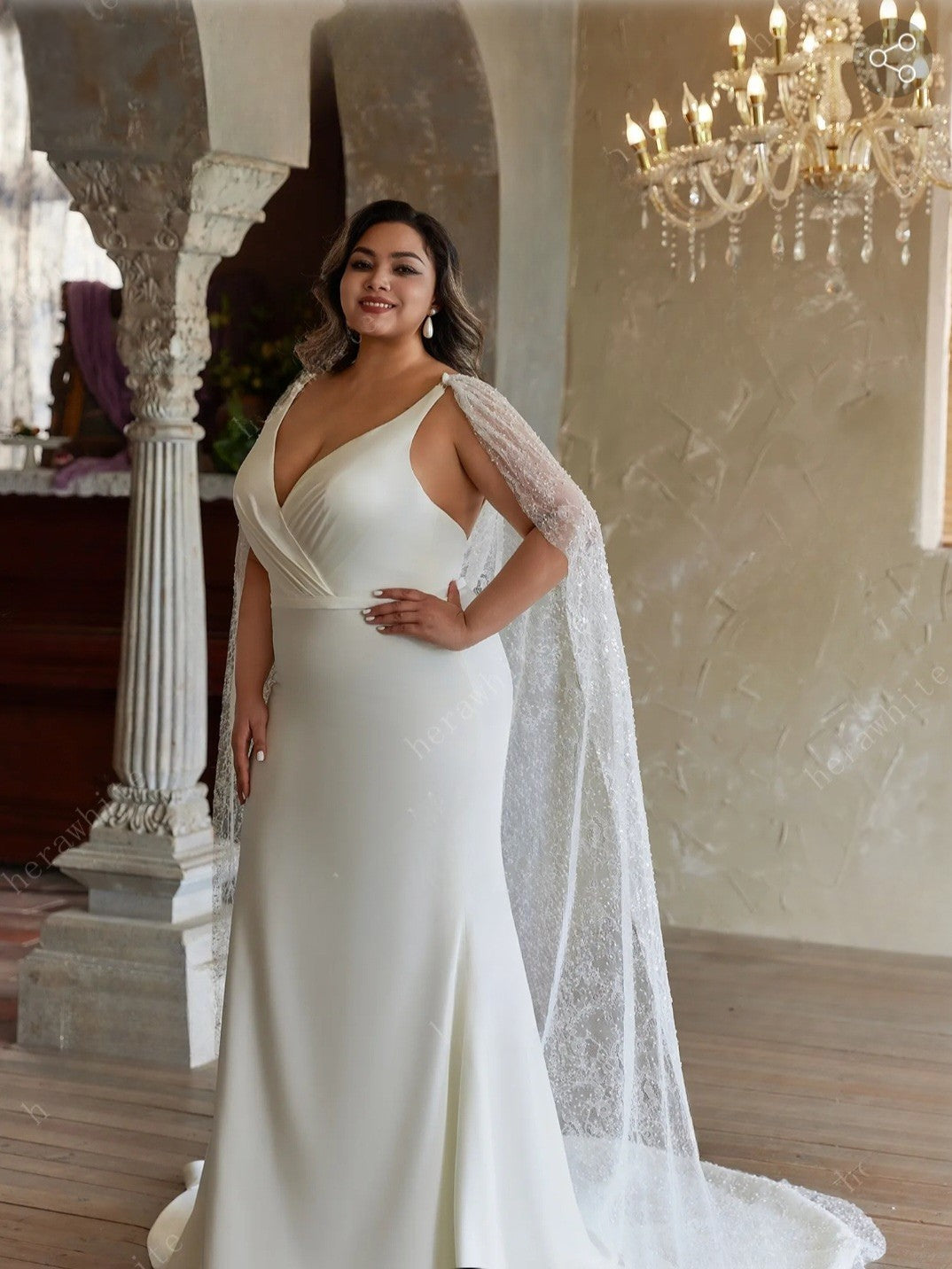 Woman in a white wedding dress with a lace cape standing in an elegant indoor setting.