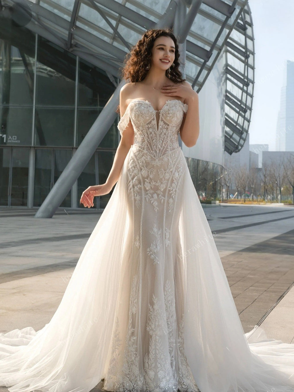 Woman in a white lace wedding dress standing in front of modern architectural structures.
