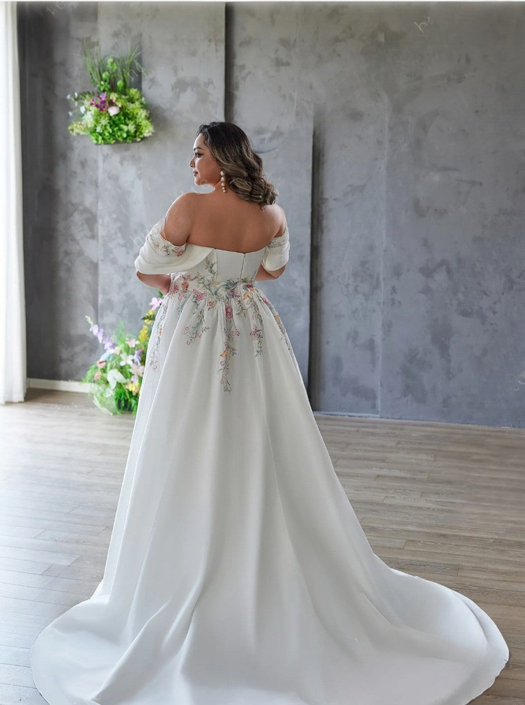 Woman in a white wedding dress with floral embroidery standing in a room with gray walls and wooden floor.