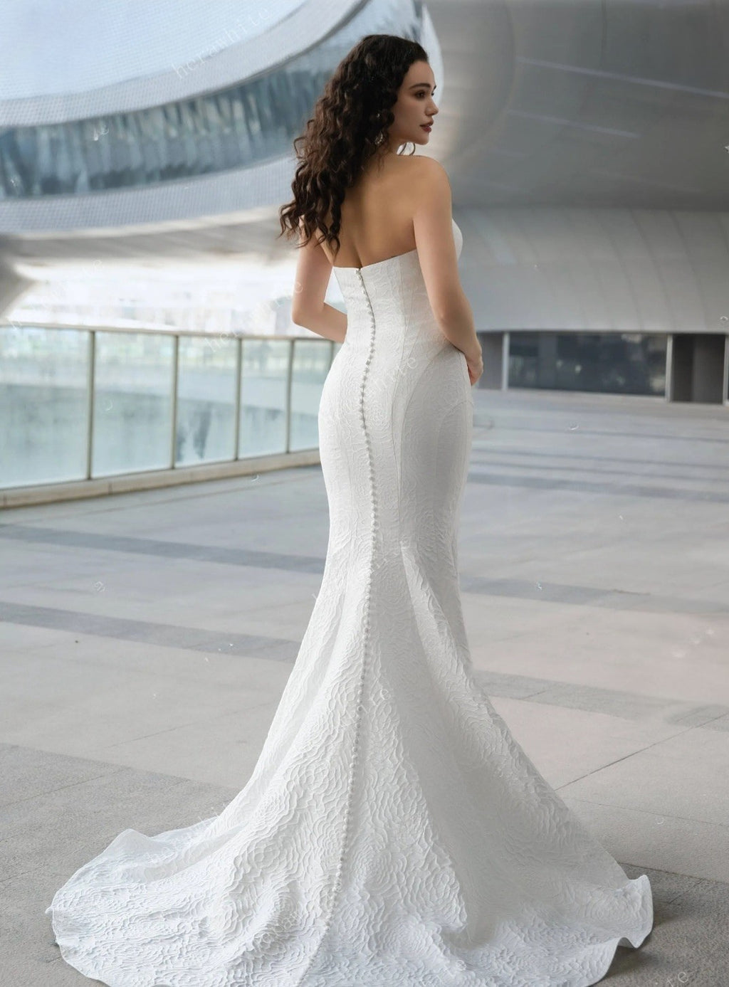 Woman in a white strapless wedding dress standing in an indoor setting with large windows.