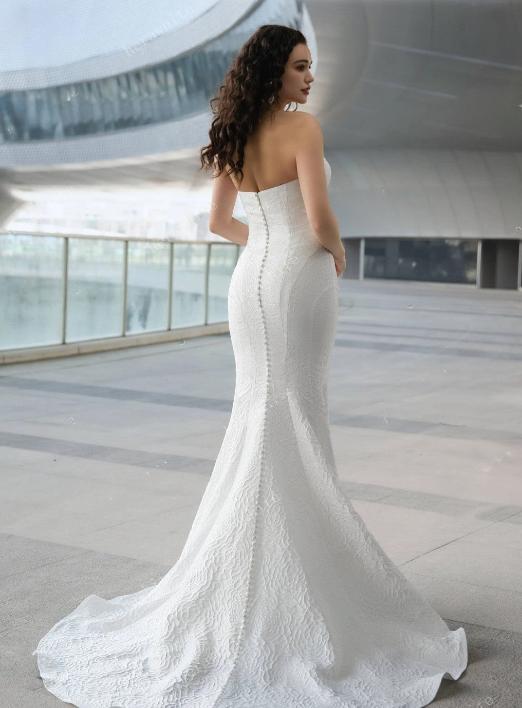 Woman in a white strapless wedding dress standing in an indoor setting with large windows.